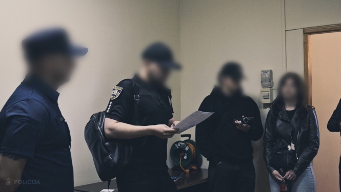 Valsts policijas darbinieki iekštelpās ar dokumentu rokās un vairāki cilvēki ar aizklātām sejām stāv blakus