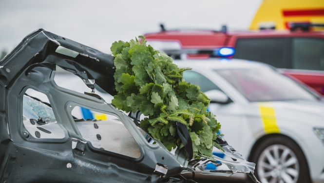 Bojātas automašīnas priekšdaļa ar Jāņu vainagu pēc ceļu satiksmes negadījuma; fonā redzams policijas un operatīvais transports ar ieslēgtām bākugunīm