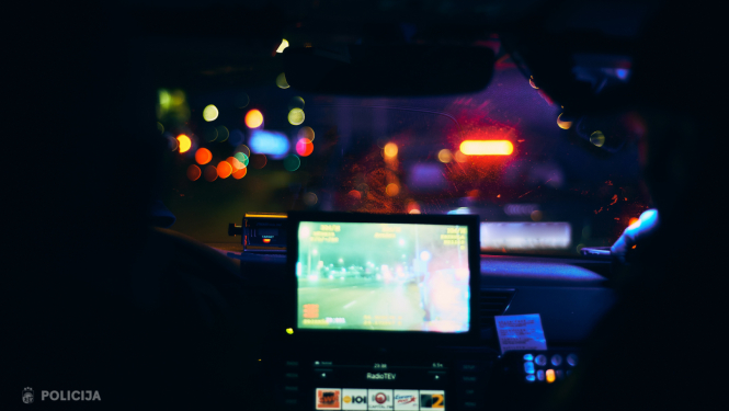 Skats no policijas automašīnas salona nakts laikā; caur priekšējo stiklu redzamas pilsētas gaismas un priekšā braucošs transportlīdzeklis, bet panelī redzams darba aprīkojuma ekrāns