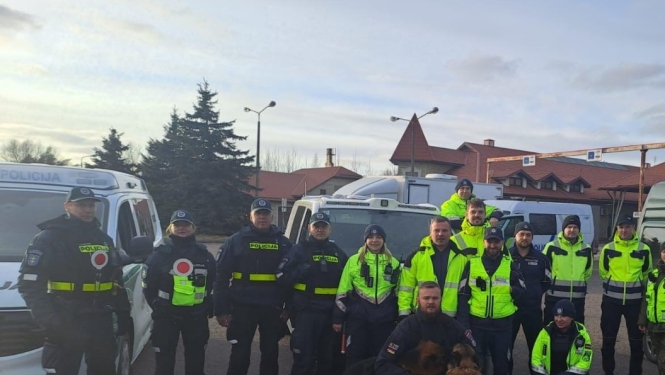 Valsts policijas un robežsardzes darbinieku grupa kopīgā foto stāv starp dienesta transportlīdzekļiem, priekšplānā redzami divi kinologi ar dienesta suņiem