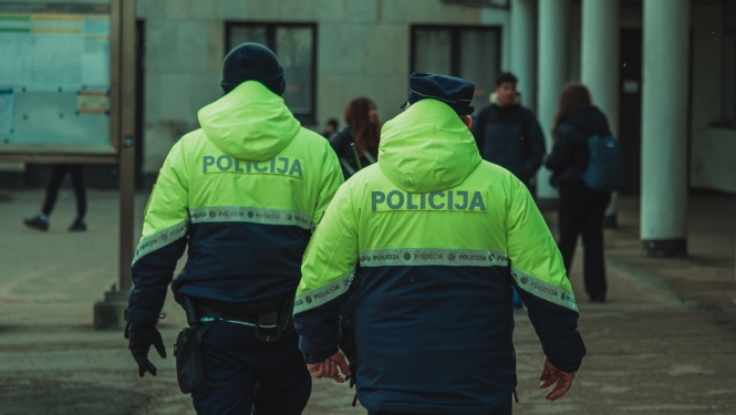 Divi Valsts policijas darbinieki koši dzeltenzaļās formas jakās dodas pa āra teritoriju pie ēkas. Fonā redzami cilvēki un pilsētvides elementi