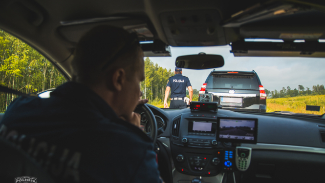 Skats policijas mašīnas iekšienē, pie stūres policists dienesta formā