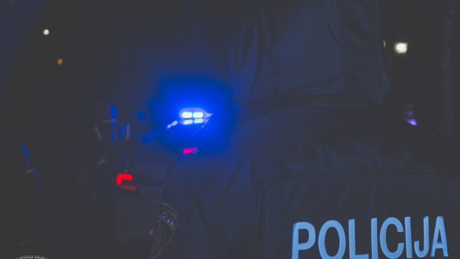Tumsā redzama policijas automašīnas zilā bākuguns un policists no mugurpuses, uz formas tērpa uzraksts POLICIJA