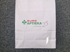 Balts papīra iepirkumu maisiņš ar uzrakstu ‘EURO APTIEKA’ novietots uz pelēkas virsmas.