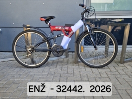 Sarkans un melns velosipēds ar priekšējo amortizācijas dakšu, aizmugurējo amortizatoru, dubļu sargiem, bagāžnieku un melnu sēdekli novietots velonovietnē pie ēkas.