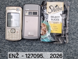 Divi mobilie telefoni un kaķu barības paciņa novietoti blakus uz pelēka paklāja. Kreisajā pusē atrodas gaišas krāsas Nokia mobilais tālrunis ar ciparu tastatūru, vidū – tumšpelēks mobilais tālrunis ar kameru, bet labajā pusē – atvērta “Sheba Select Slices” kaķu barības paciņa ar vistas gaļu.
