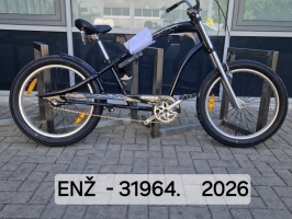 Melns kruīza tipa velosipēds ar augstu stūri, plata izmēra riepām ar baltiem sāniem, dubļu sargiem un melnu sēdekli novietots velonovietnē pie ēkas.