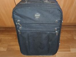Melns Travelite auduma čemodāns uz riteņiem ar priekšējām kabatām novietots uz koka grīdas