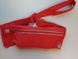 Sarkana jostas soma ar regulējamu sarkanu jostu un plastmasas aizdari. Somas priekšpusē ir horizontāla kabata ar rāvējslēdzēju un atstarojošu joslu, bet apakšējā labajā stūrī redzams apaļš “lululemon” logotips. Soma novietota uz baltas virsmas.