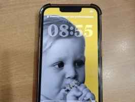 Apple mobilais telefons caurspīdīgā vāciņā fotografēts no priekšpuses, ar ieslēgtu ekrānu un no aizmugures uz gaišas koka virsmas