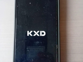 Melns skārienjutīgs mobilais telefons ar ieslēgtu ekrānu, uz kura redzams “KXD” logotips. Ekrānam redzami bojājumi un krāsaini artefakti labajā apakšējā stūrī. Telefons novietots uz gaišas virsmas.
