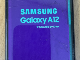 Samsung Galaxy A12 mobilais telefons ar redzamiem ekrāna bojājumiem un ieslēgtu displeju uz gaišas koka virsmas.