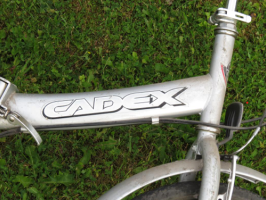 Saliekamais velosipēds "CADEX CLASSIC 110" gaiši pelēkā krāsā, un melnu metāla grozu uz bagāžas nodalījuma