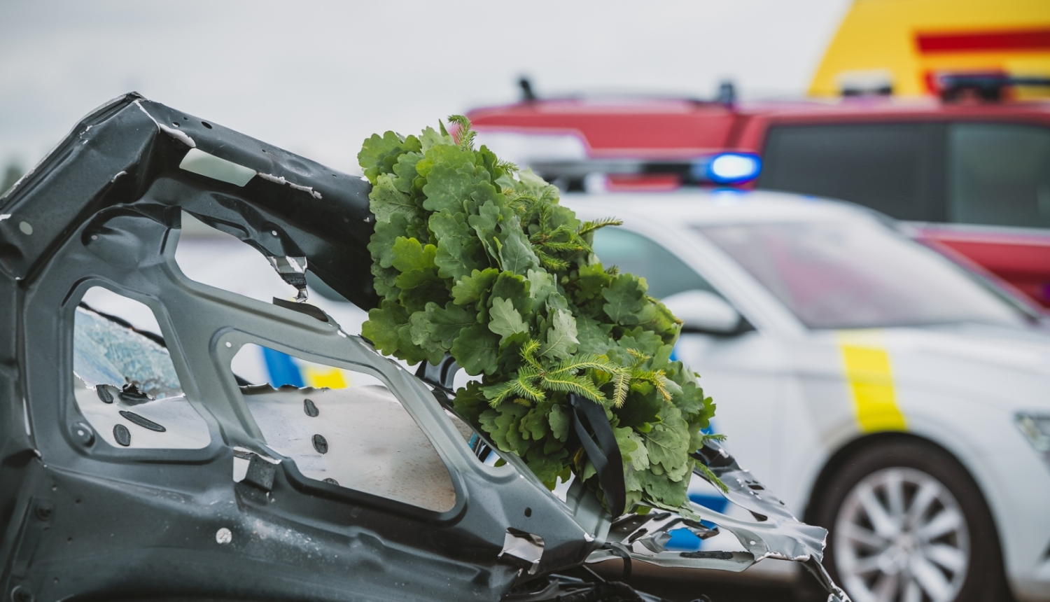 Bojātas automašīnas priekšdaļa ar Jāņu vainagu pēc ceļu satiksmes negadījuma; fonā redzams policijas un operatīvais transports ar ieslēgtām bākugunīm