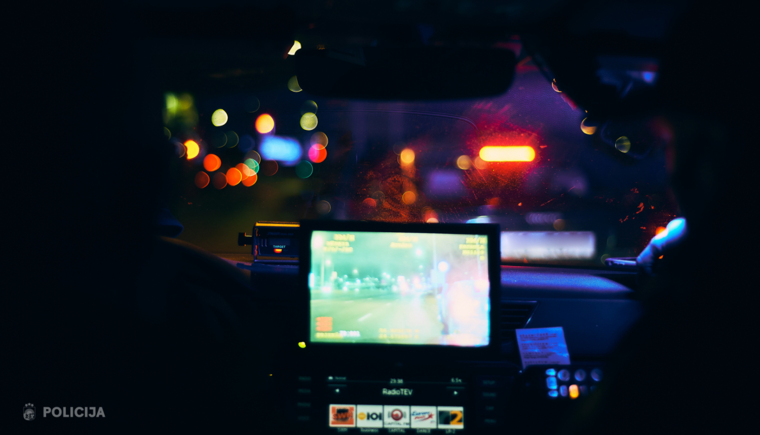 Skats no policijas automašīnas salona nakts laikā; caur priekšējo stiklu redzamas pilsētas gaismas un priekšā braucošs transportlīdzeklis, bet panelī redzams darba aprīkojuma ekrāns