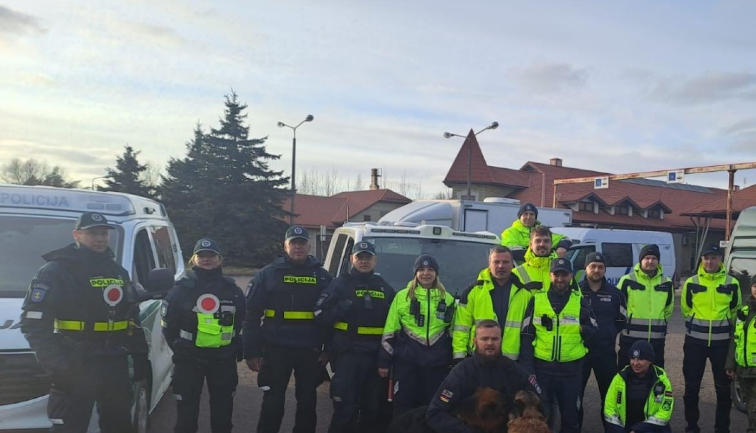 Valsts policijas un robežsardzes darbinieku grupa kopīgā foto stāv starp dienesta transportlīdzekļiem, priekšplānā redzami divi kinologi ar dienesta suņiem