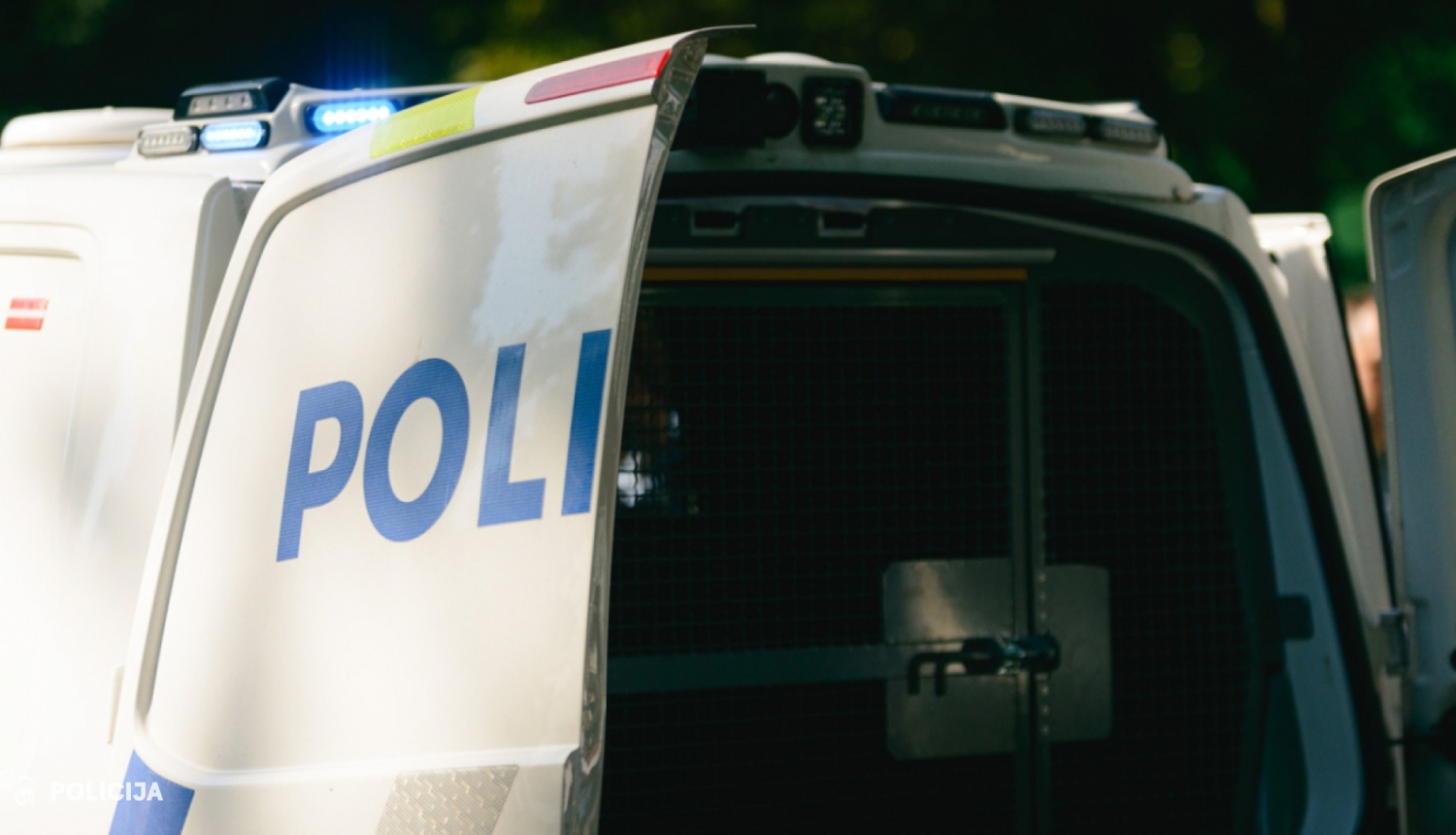 Atvērtas Valsts policijas transportlīdzekļa aizmugurējās durvis ar redzamu uzrakstu “POLICIJA” un ieslēgtām zilajām bākugunīm