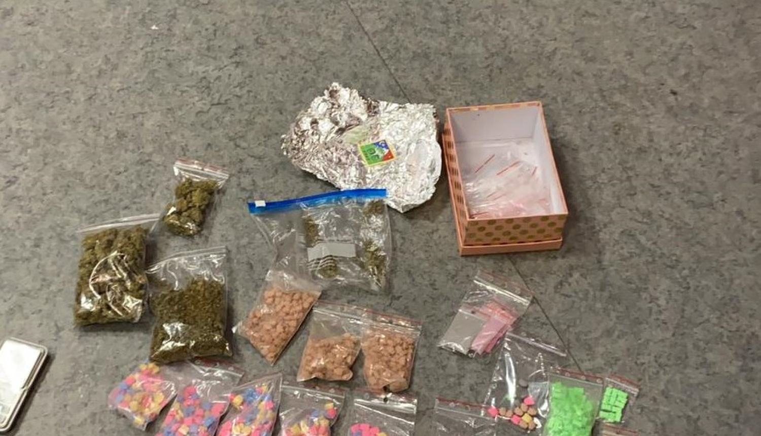 Uz grīdas nolikti vairāki caurspīdīgi plastmasas maisiņi ar dažādu krāsu tabletēm - narkotikām, marihuānu, folijā ietītas markas, pelēks mobilais tālrunis