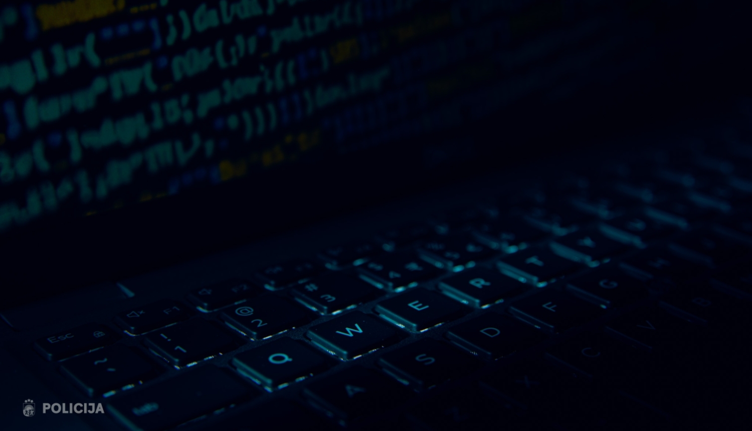 Tumšā, zilganā apgaismojumā tuvplānā redzama datora tastatūra, bet fonā izplūduši programmēšanas koda fragmenti