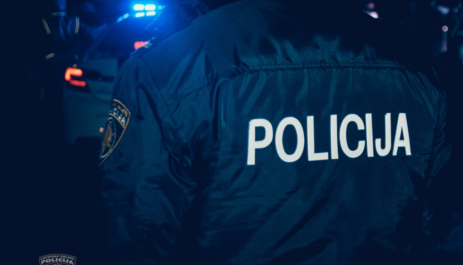 Policists formā, uz muguras uzrakstīts Policija, tālumā fonā policijas mašīna ar ieslēgtām bākugunīm, tumšajā diennakts laikā
