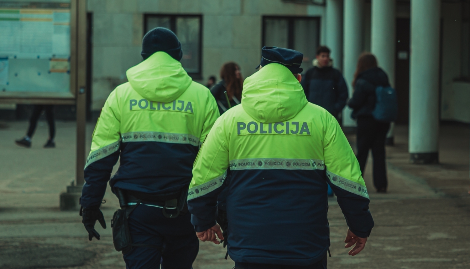 Divi Valsts policijas darbinieki koši dzeltenzaļās formas jakās dodas pa āra teritoriju pie ēkas. Fonā redzami cilvēki un pilsētvides elementi