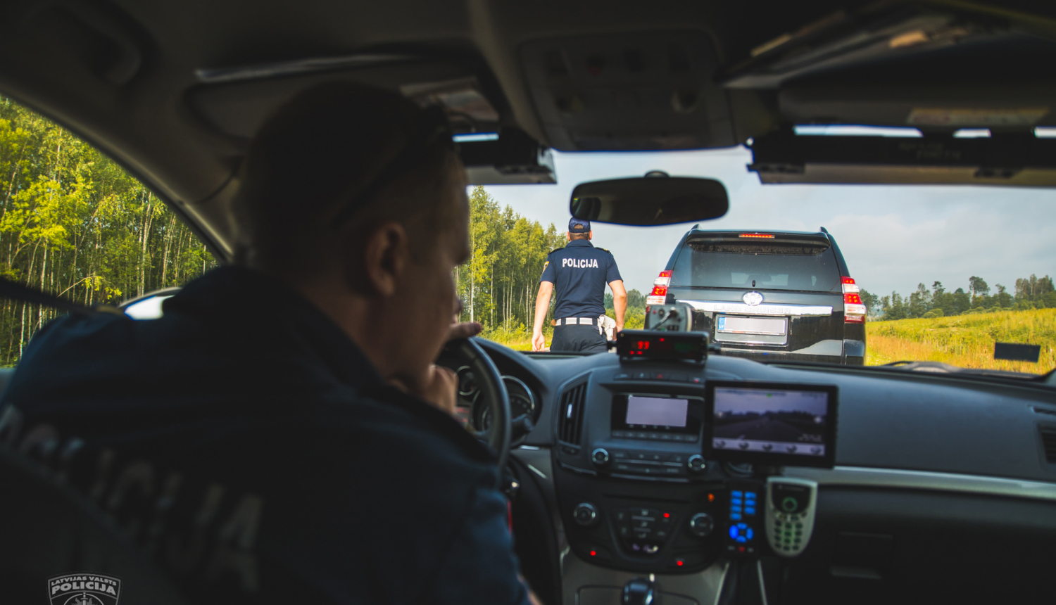 Skats policijas mašīnas iekšienē, pie stūres policists dienesta formā