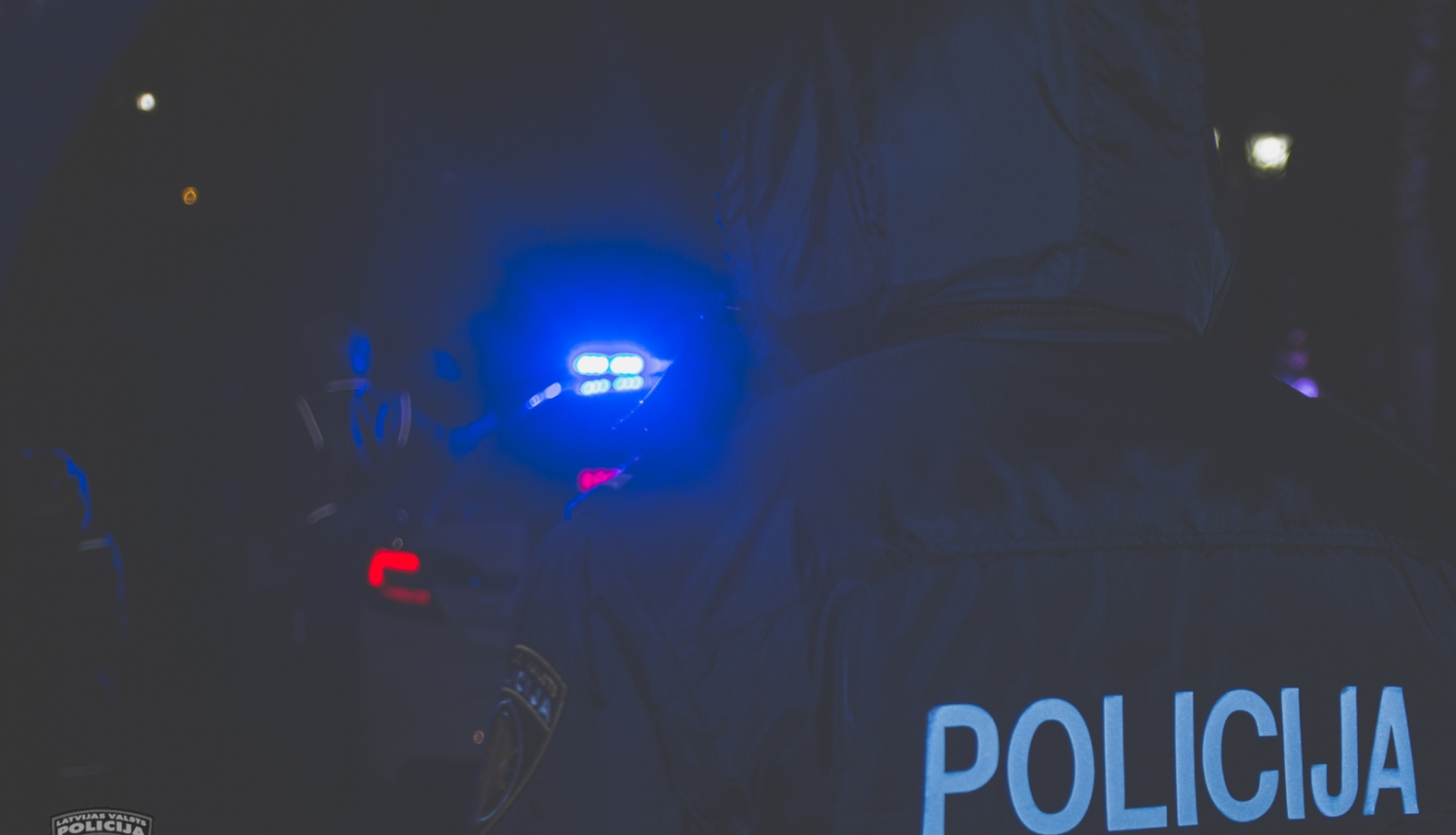 Tumsā redzama policijas automašīnas zilā bākuguns un policists no mugurpuses, uz formas tērpa uzraksts POLICIJA