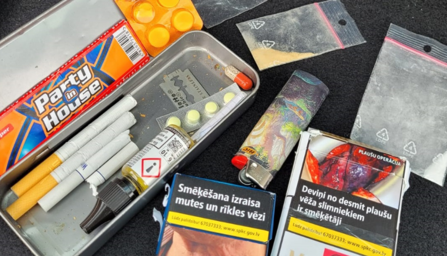 Metāla penālis, kurā atrodas cigaretes, tabletes, žilete, pie penāļa cigarešu paciņas, caurspīdīgi plastikāta maisiņi ar baltas un oranžas krāsas pulverīšiem