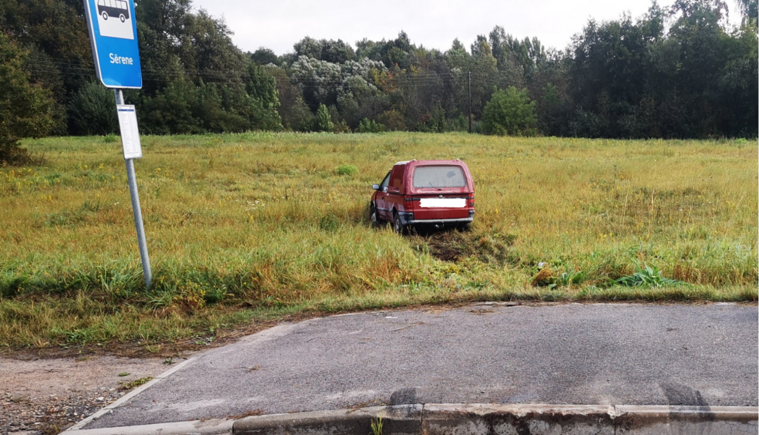 Sarkana automašīna iebraukusi pļavā, priekšplānā autobusa pietura