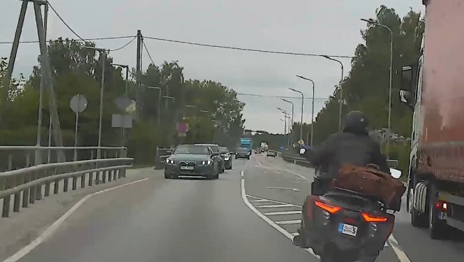 Ceļš ar vairākām braukšanas joslām, priekšplānā pa labo pusi brauc motociklists, blakus kravas automašīnai, bet pretējā virzienā pārvietojas vieglās automašīnas