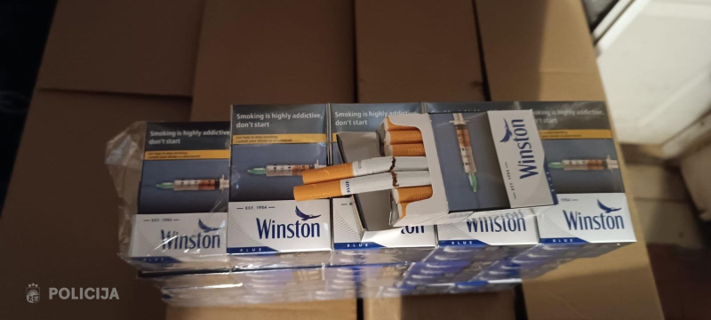 Uz kartona kastēm sakrautas vairākas cigarešu paciņas “Winston”, dažas no tām atvērtas ar redzamām cigaretēm.