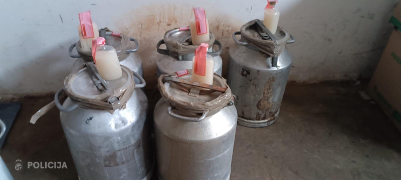 Vairākas metāla kannas ar nelegāli pildītiem plastmasas traukiem, kas, iespējams, satur nezināmas izcelsmes alkoholu, novietotas telpā uz grīdas.