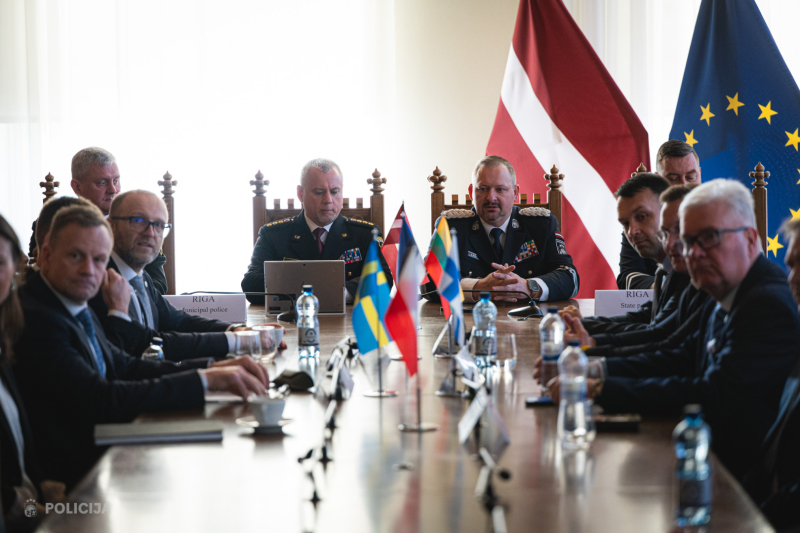 Vairāku valstu un institūciju pārstāvji sēž pie gara konferenču galda, daļai esot formastērpos, daļai lietišķā apģērbā. Galda centrā izvietoti vairāku valstu karodziņi, bet fonā redzami Latvijas un Eiropas Savienības karogi