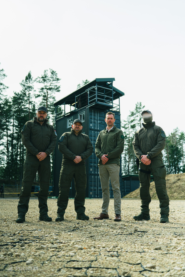 Četri vīrieši stāv āra apmācību teritorijā pie taktiskās mācību būves mežainā apvidū