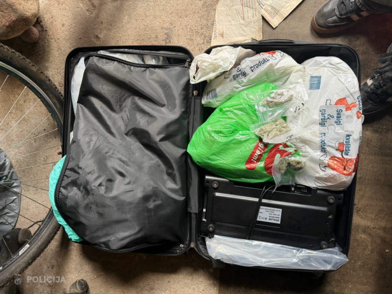 Atvērts melns čemodāns uz grīdas, kurā redzami vairāki plastmasas maisi un iepakojumi ar zaļganas augu izcelsmes vielu