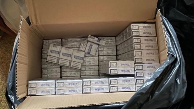 Atvērtā kartona kastē redzamas daudzas baltas cigarešu paciņas