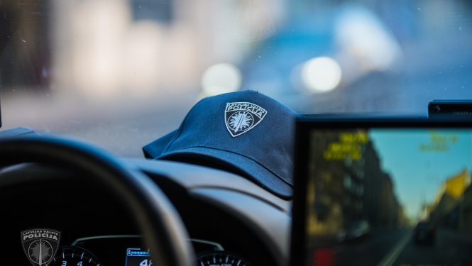 Policijas automašīnas iekšpuse, redzama stūre, daļa no ekrāna, uz priekšējā paneļa uzlikta policista cepure