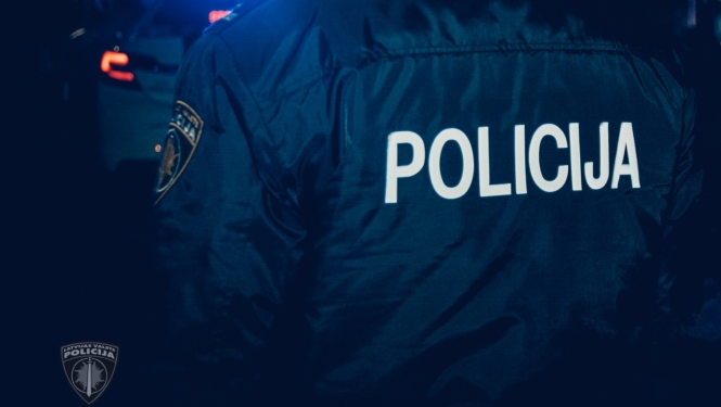 Policists formā, uz muguras uzrakstīts Policija, tālumā fonā policijas mašīna ar ieslēgtām bākugunīm, tumšajā diennakts laikā