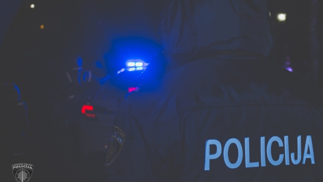 Tumsā redzama policijas automašīnas zilā bākuguns un policists no mugurpuses, uz formas tērpa uzraksts POLICIJA