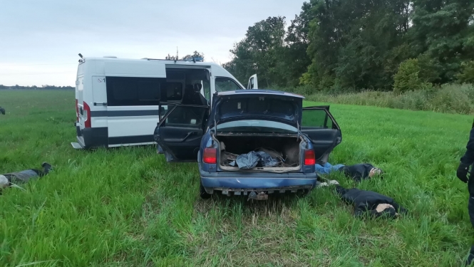 Policijas automašīna piebraukusi priekšā vieglam auto, kam visas durvis atvērtas, arī bagāžas nodalījums. Blakus automašīnai zālē guļ 3 vīrieši ar seju pret zemi