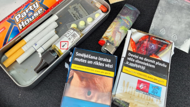 Metāla penālis, kurā atrodas cigaretes, tabletes, žilete, pie penāļa cigarešu paciņas, caurspīdīgi plastikāta maisiņi ar baltas un oranžas krāsas pulverīšiem