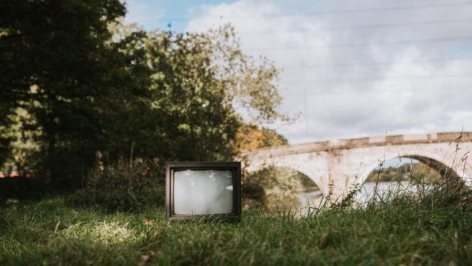 Vecs televizors novietots zālē, fonā redzami krūmi un akmens tilts ar arkām; saulaina diena; vasara