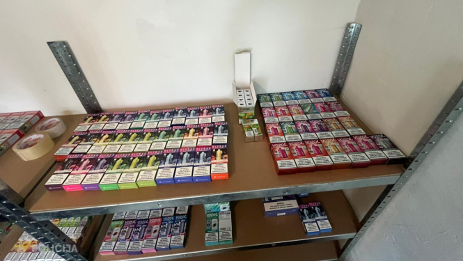 Metāla plauktā novietotas daudzas elektronisko cigarešu un veipu iepakojumu kastes dažādās krāsās, sakārtotas vairākās rindās uz diviem plauktiem