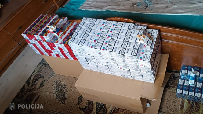 Istabā pie gultas uz kartona kastēm sakrauts liels daudzums cigarešu paciņu dažādos iepakojumos; daļa paciņu novietota arī atsevišķi uz grīdas