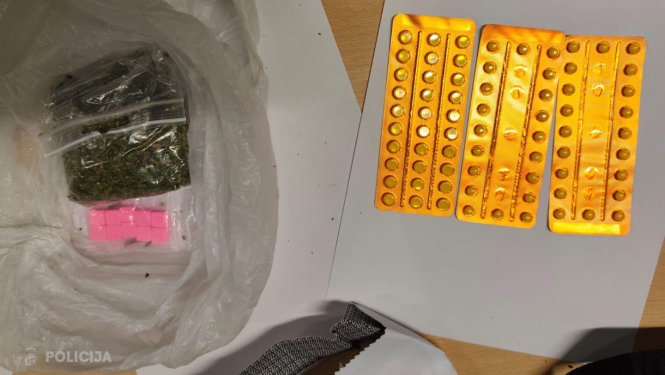 Uz galda stāv plastmasas maisiņš ar vairākiem nelieliem iepakojumiem, kuros ir zaļas augu izcelsmes vielas un rozā tabletes, blakus novietotas plāksnītes ar tabletēm