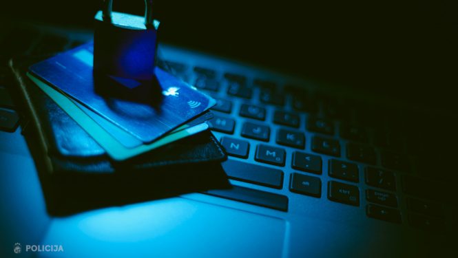 Tumšā zilganā apgaismojumā uz klēpjdatora tastatūras novietots maks ar bankas kartēm, uz kurām uzlikta piekaramā slēdzene, simboliski attēlojot finanšu un datu drošību.