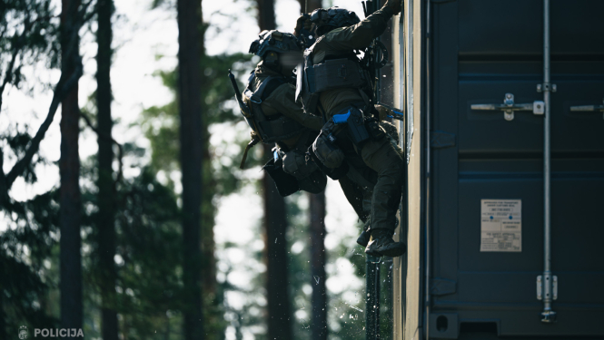 Divi Valsts policijas specvienības darbinieki taktiskajā ekipējumā treniņa laikā iekļūst ēkā mežainā apvidū
