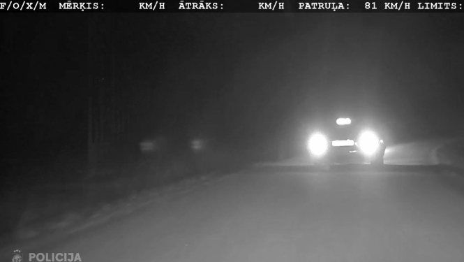Melnbalts nakts redzamības kameras kadrs uz ceļa, kur pretī brauc automašīna ar spilgtiem lukturiem