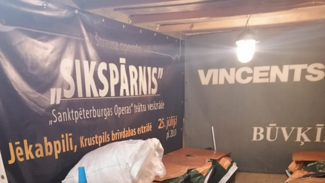 Zilas plastmasas mucas, pārsegtas ar finiera gabaliem, priekšplānā alumīnija kanna, fonā pie sienas operas izrādes "Sikspārnis" platāts un uzņēmuma "Vincents" reklāma  