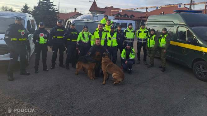 Valsts policijas un robežsardzes darbinieku grupa kopīgā foto stāv starp dienesta transportlīdzekļiem, priekšplānā redzami divi kinologi ar dienesta suņiem